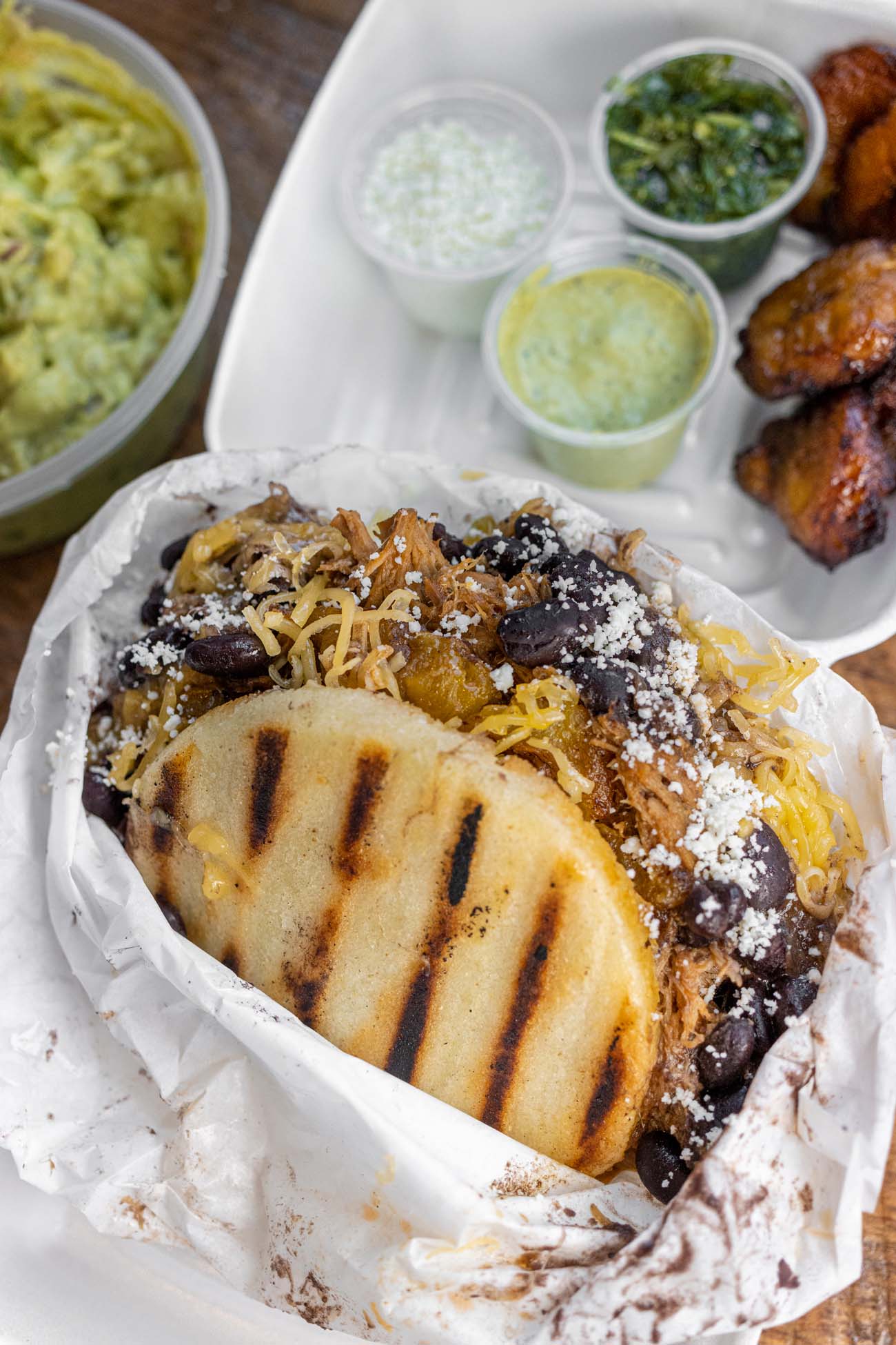 Close-up overhead view of a Venezuelan arepa in a paper-lined container from Chellas Arepa Kitchen. The grilled cornmeal pocket is filled with shredded meat, black beans, and cheese, topped with a sprinkling of cotija cheese. Surrounding the arepa are various accompaniments: a large container of fresh guacamole on the left, small cups of different sauces (likely including chimichurri and their signature Mad Llama hot sauce), and fried sweet plantains on the right. The image showcases the generous portions and vibrant colors of the authentic South American cuisine offered at this downtown Lancaster hidden gem.