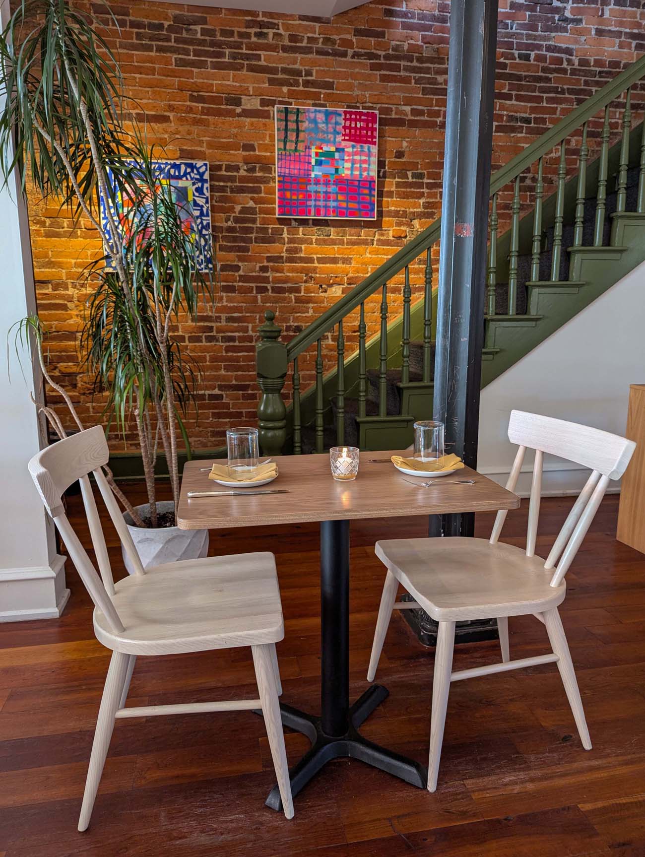 A cozy dining area at Passerine Lancaster featuring a two-person wooden table with light wood chairs, exposed brick walls decorated with colorful geometric artwork, a green painted staircase with traditional spindles, potted plants, and warm ambient lighting