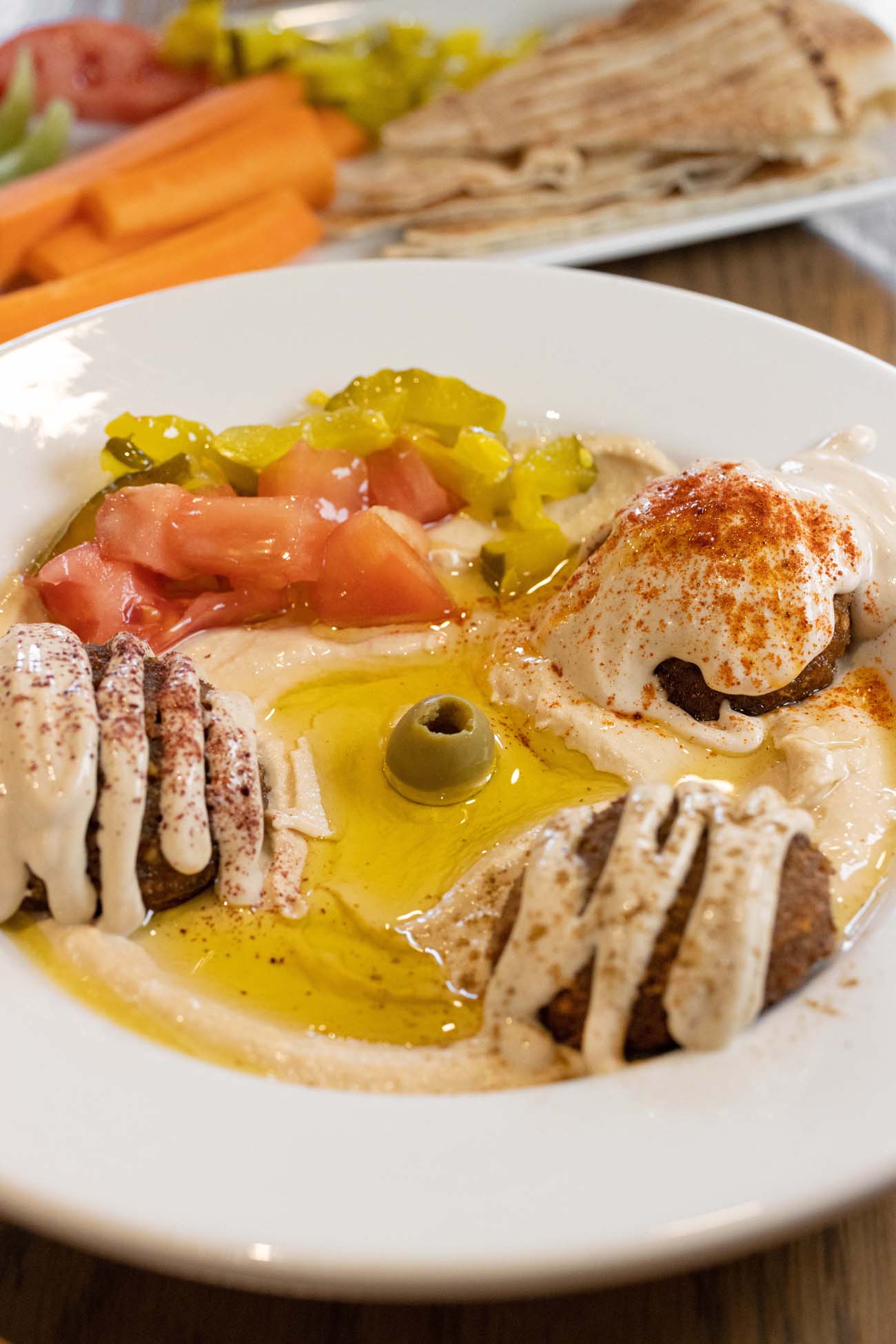 Hummus Bowl with falafel served with pita and veggies on the side from Layali El Sham. A close-up of a Mediterranean mezze platter served in a white bowl. The base appears to be creamy hummus with golden olive oil and topped with fresh diced tomatoes, pickled yellow peppers, and a single green olive in the center. The dish is dusted with paprika or sumac for color and flavor. Around the edges are small falafel with a white sauce drizzled over them. In the background slightly out of focus, there's a platter with sliced vegetables including carrots and other crudités. This appetizer represents the diverse international cuisine available in downtown Lancaster's restaurant scene, showcasing Mediterranean influences with fresh ingredients and traditional presentation.
