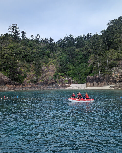 View of a beach in the Whitsunday Islands from a boat just off the shore. In the water, a red dinghy holds snorkellers.